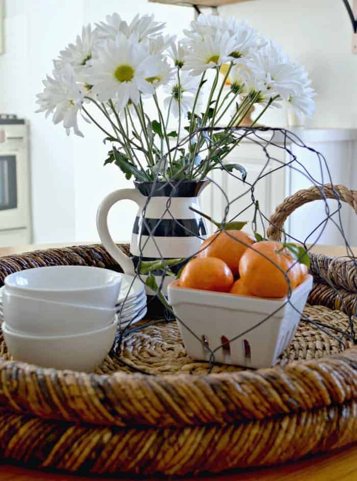 woven tray with white dishes, container of oranges and pitcher of daisies