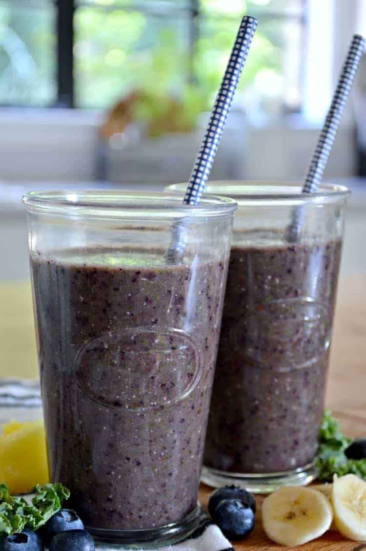 2 kale blueberry smoothies with a blue straw sitting on a wooden counter