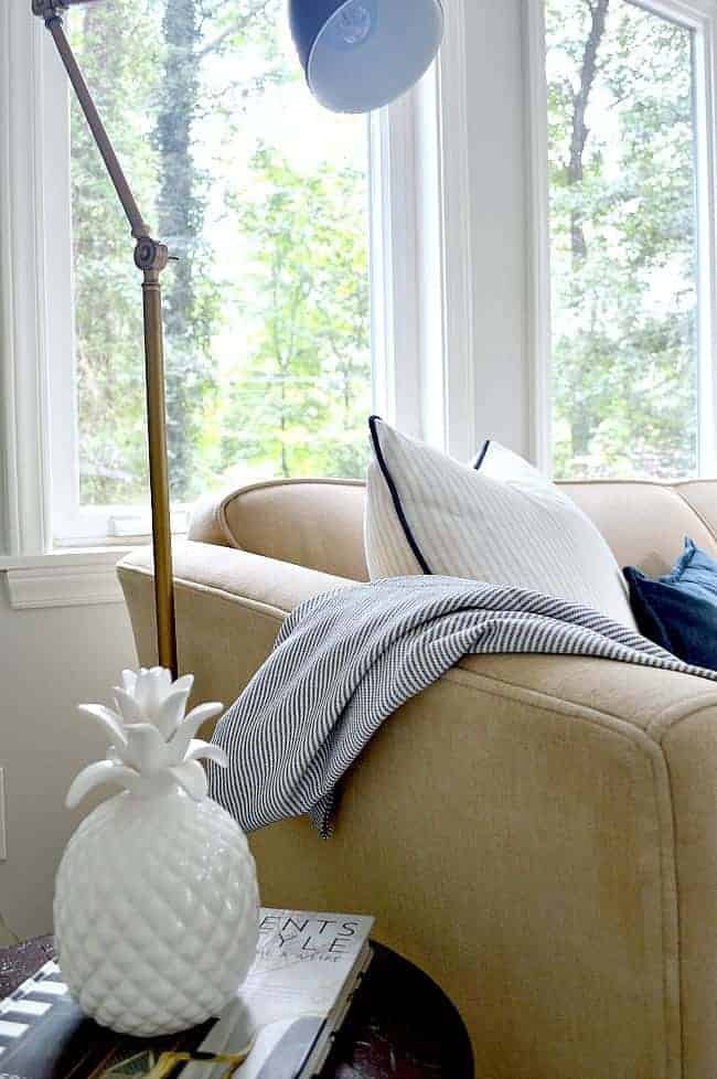 corner of beige sofa with floor lamp and stool with a book and ceramic pineapple on it.