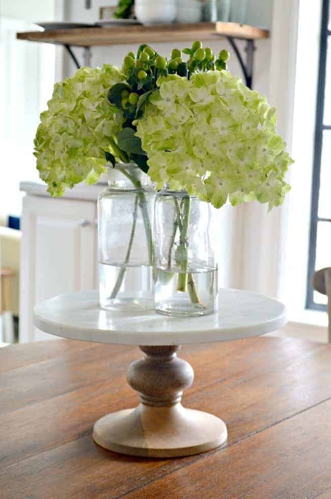 3 glass bottles with hydrangea on a marble cake stand in the middle of a wood table