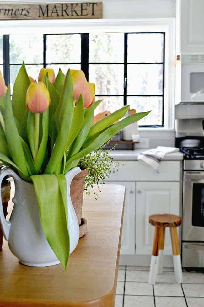 A pitcher of yellow and orange tulips on a butcher block island with kitchen window and wooden stool in the background