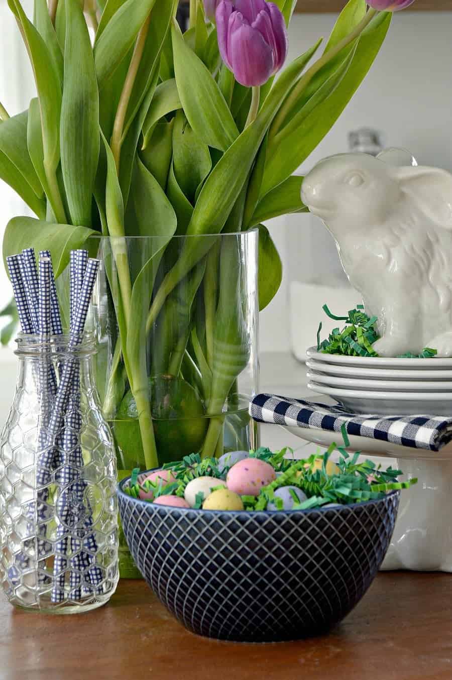 spring vignette with purple tulips, white bunnies, a blue checked napkin, blue straws in a glass bottle and a navy bowl with candy eggs 