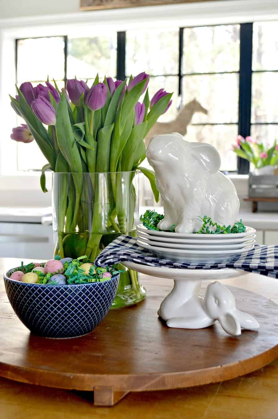 spring vignette with purple tulips, white bunnies, a navy bowl with candy eggs on a large wood cutting board