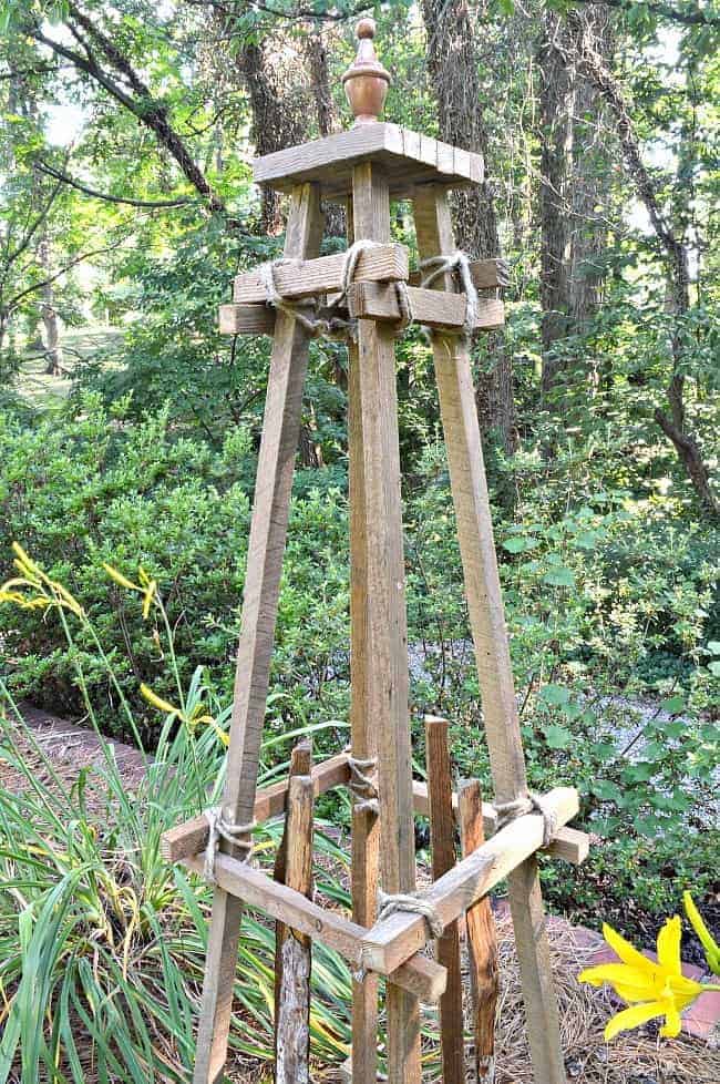 top half of a wooden garden obelisk sitting in the middle of a garden with daylilies