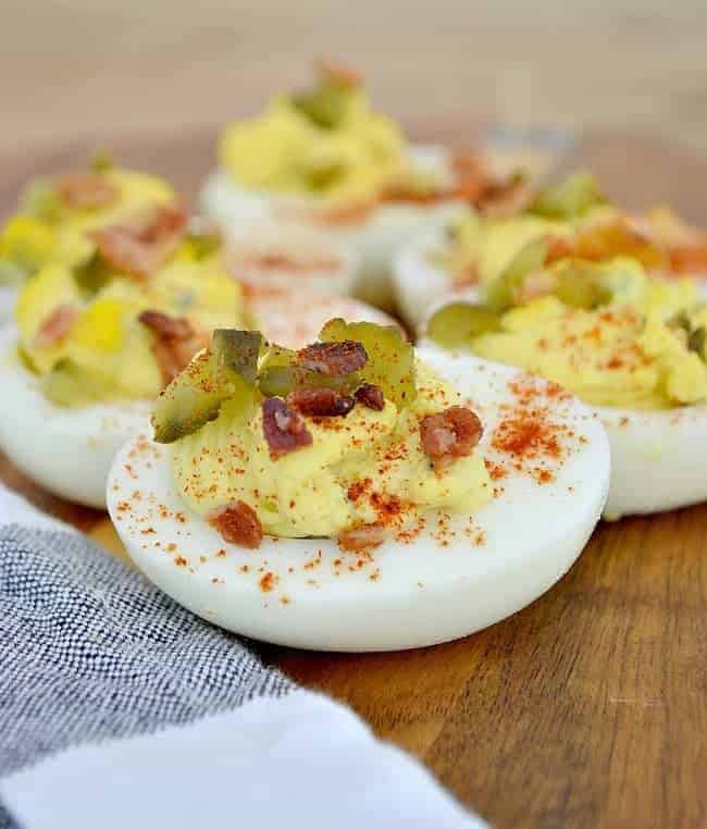 deviled eggs on a wooden board with bacon, pickles and paprika sprinkled on top