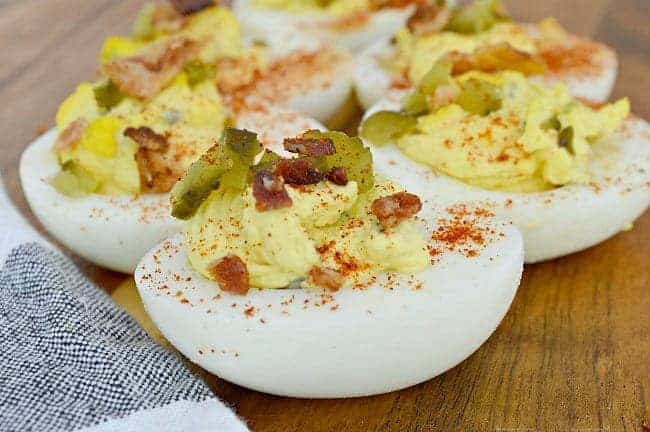 deviled egg halves with pickles and bacon on top on wood board with black and white towel in corner