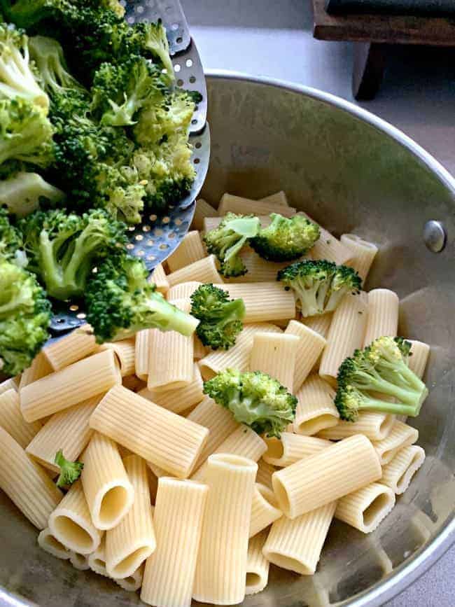 pouring steamed broccoli on to cooked noodles in pot