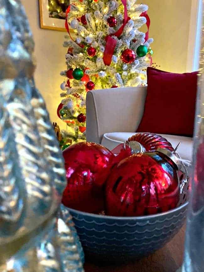 a bowl of red shiny ornaments with a flocked Christmas tree in the corner of a sunroom 