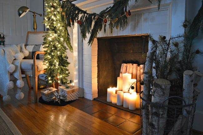 white brick fireplace with lighted candles in it and fresh greens hanging on it and a small, skinny lighted tree next to it