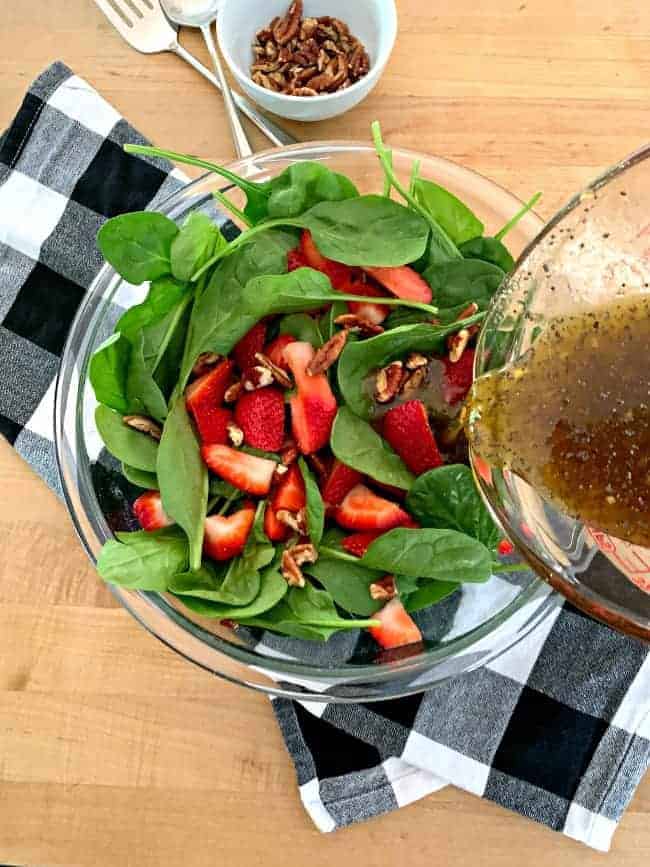 pouring poppy seed dressing over a spinach strawberry salad with chopped pecans