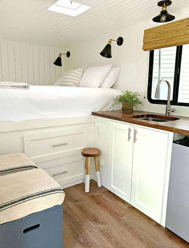 inside view of RV renovation with a big white bed, kitchen sink and counter and navy wood storage stools